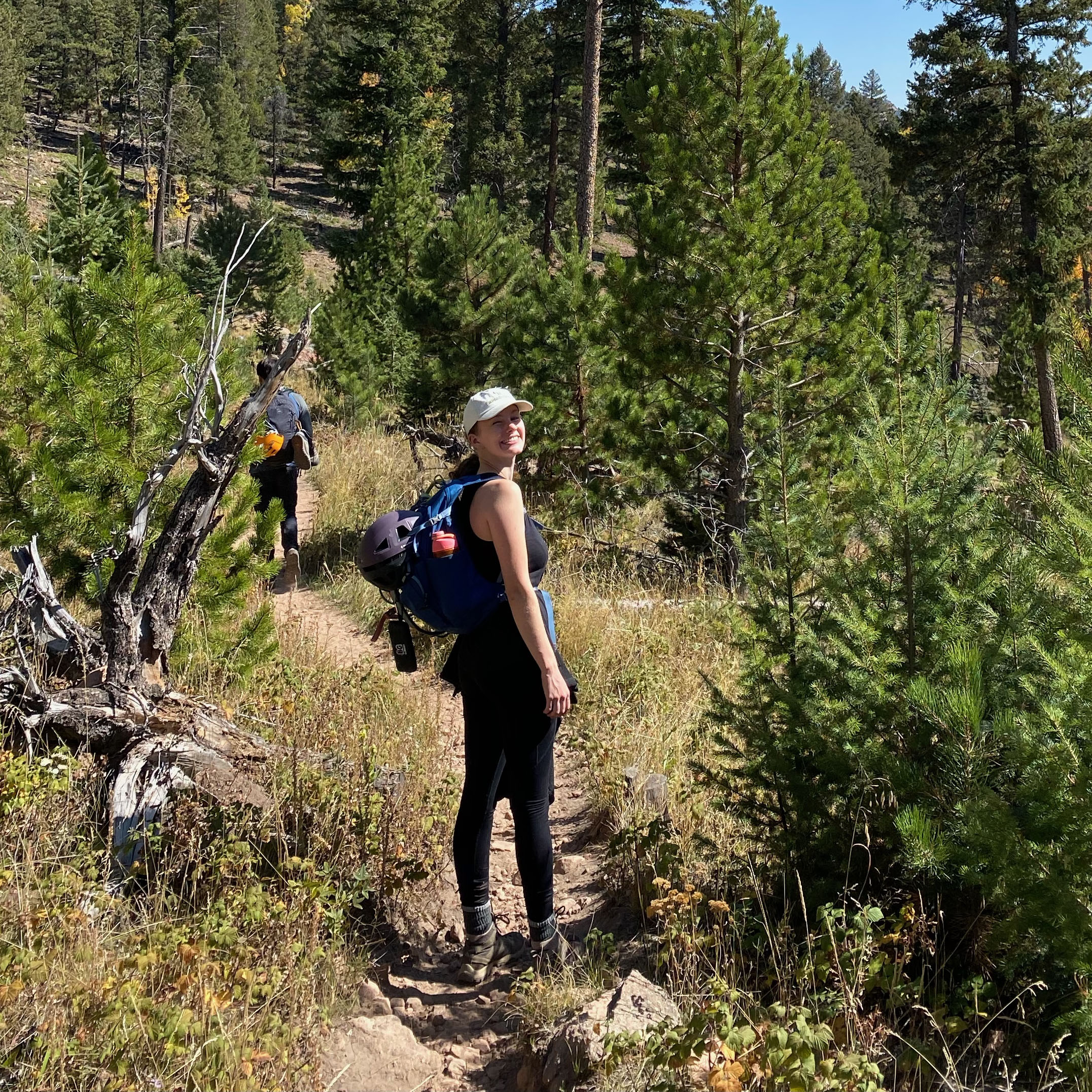 A hiker wearing a hat walking along a narrow, rocky trail through a sunlit pine forest.