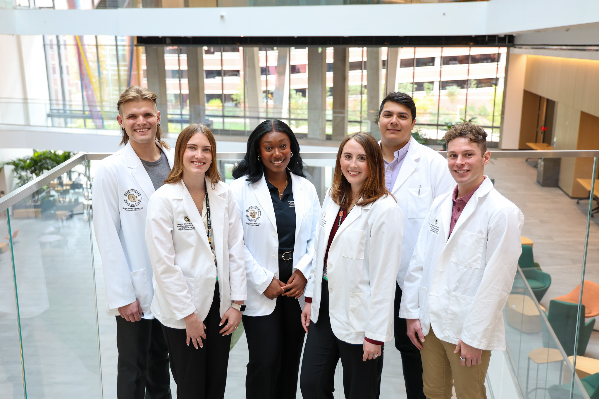 Six students wearing white lab coats pose together in a bright, modern atrium with large windows and glass railings.